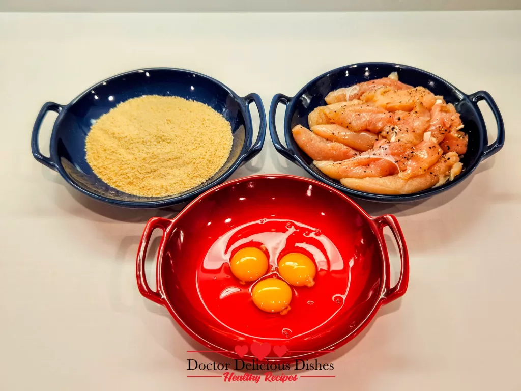 Three bowls set up for breading Crispy Air Fryer Chicken Tenders, containing eggs, seasoned breadcrumbs, and prepared chicken.