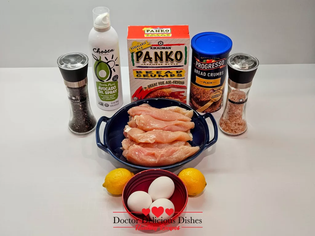 A top-down view of all ingredients needed to make Crispy Air Fryer Chicken Tenders, including chicken, breadcrumbs, eggs, seasoning, and lemons.