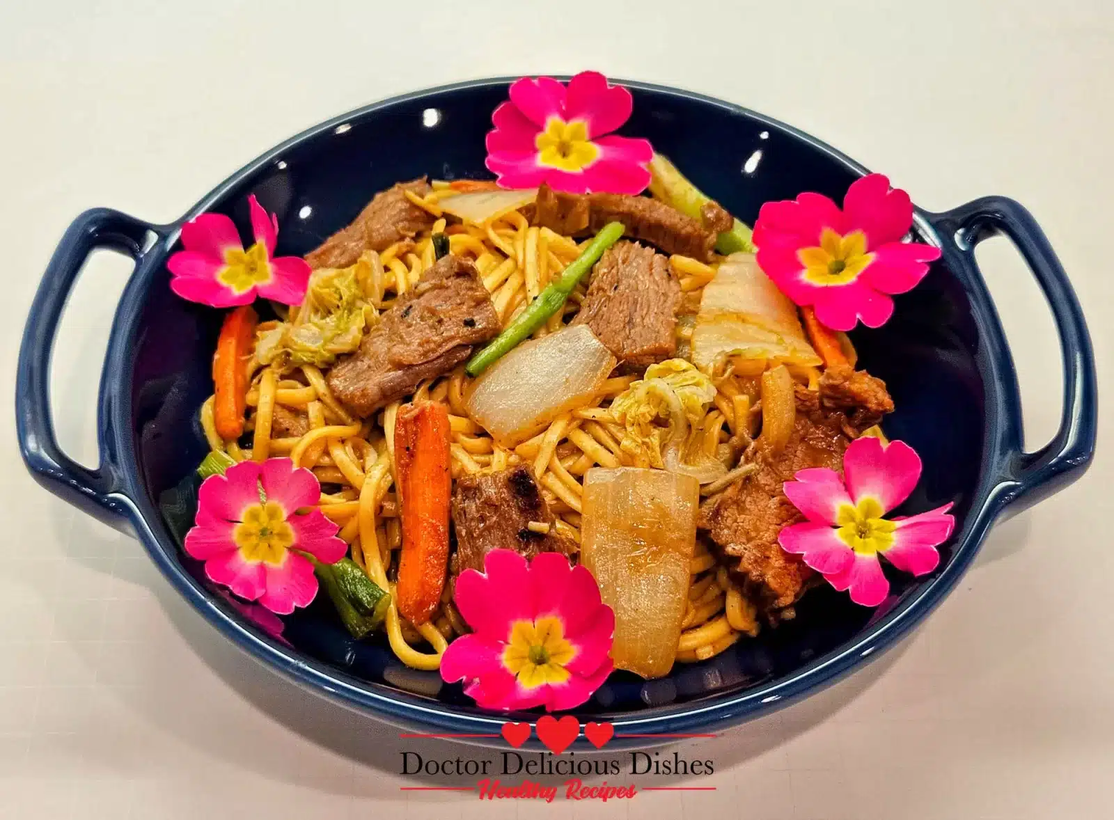 Plated Beef Lo Mein in a dark blue dish, topped with edible flowers for a vibrant presentation