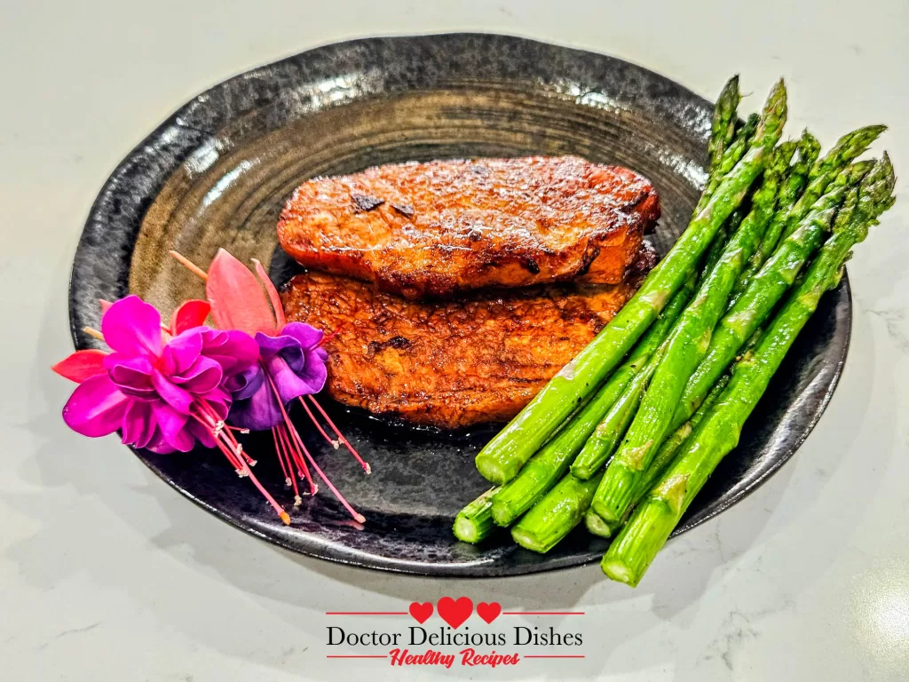 A full serving of two air fryer pork chops with bright green asparagus spears, beautifully presented on a dark, textured plate with fuchsia flowers.