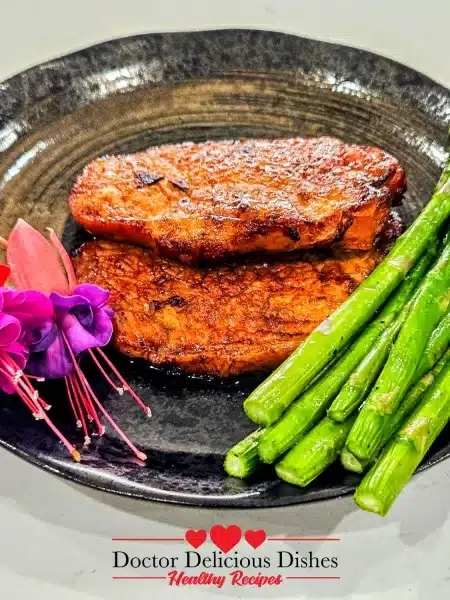 A full serving of two air fryer pork chops with bright green asparagus spears, beautifully presented on a dark, textured plate with fuchsia flowers.
