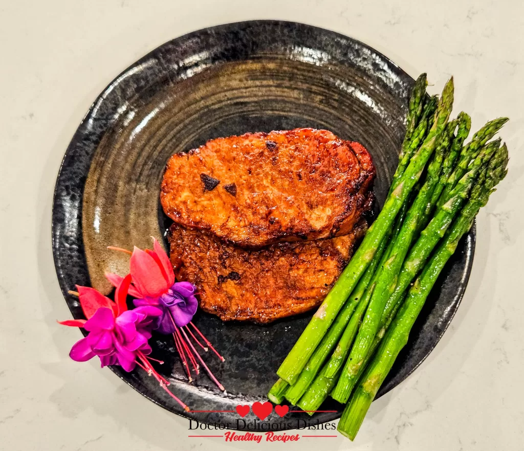 Two golden brown air-fryer pork chops and a generous serving of green asparagus, artfully arranged on a dark plate with pink and purple fuchsia flowers, ready to serve.