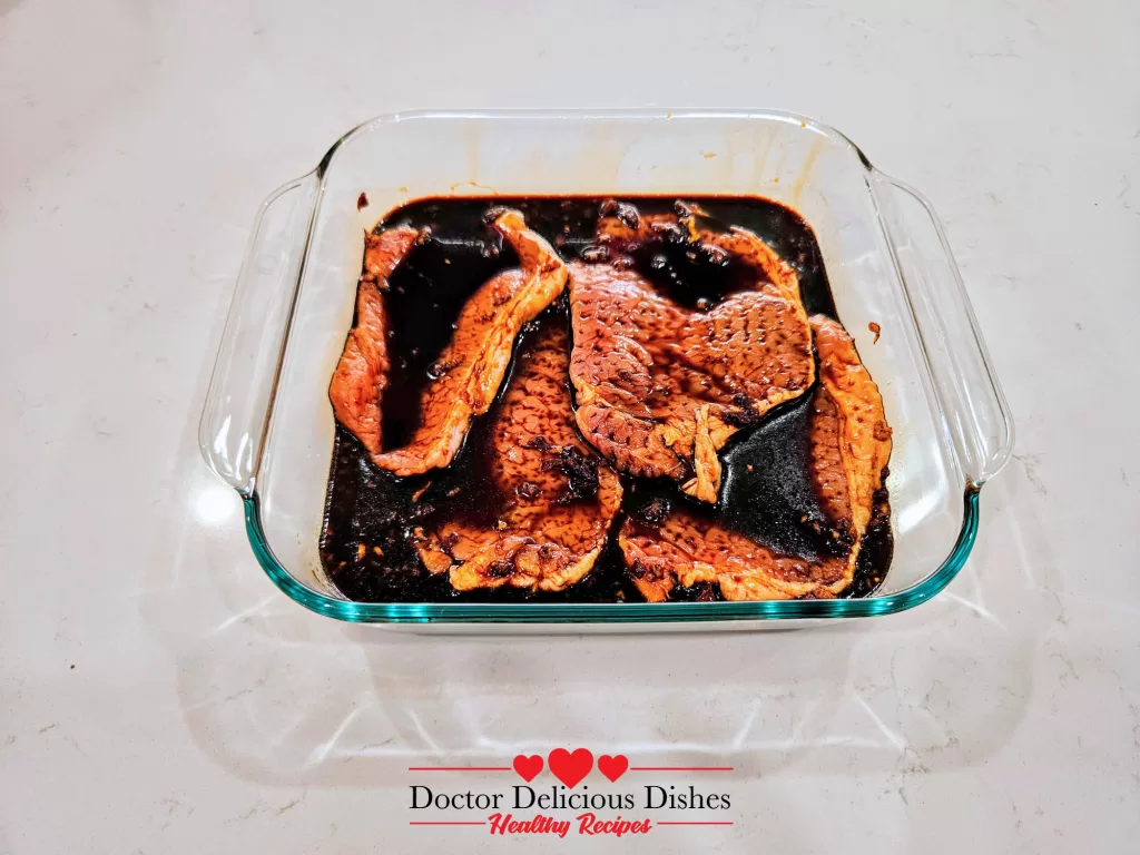 Four boneless thin cut pork chops soaking in a dark marinade in a clear glass baking dish, absorbing flavors before cooking.