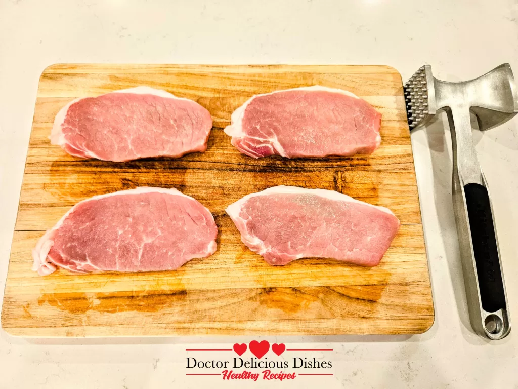 Four raw boneless thin cut pork chops neatly arranged on a wooden cutting board next to a meat tenderizer.