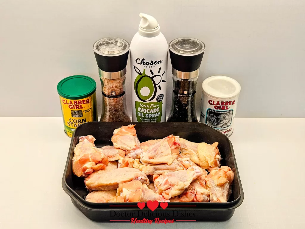 Ingredients for Air Fryer Chicken Wings including cornstarch, baking powder, avocado oil spray, and raw chicken wings in a tray.
