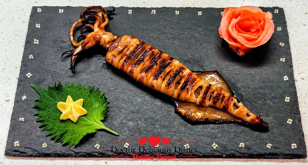 Japanese grilled squid (Ikayaki) plated on a black slate with shiso leaf, Kewpie star, and shaped pink pickled ginger garnish.