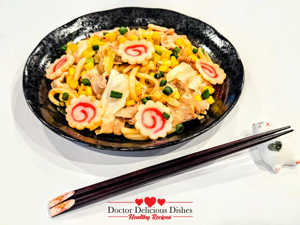 Close-up of Yaki Udon bowl with glossy noodles, pork, and narutomaki beside a white cat chopstick holder.