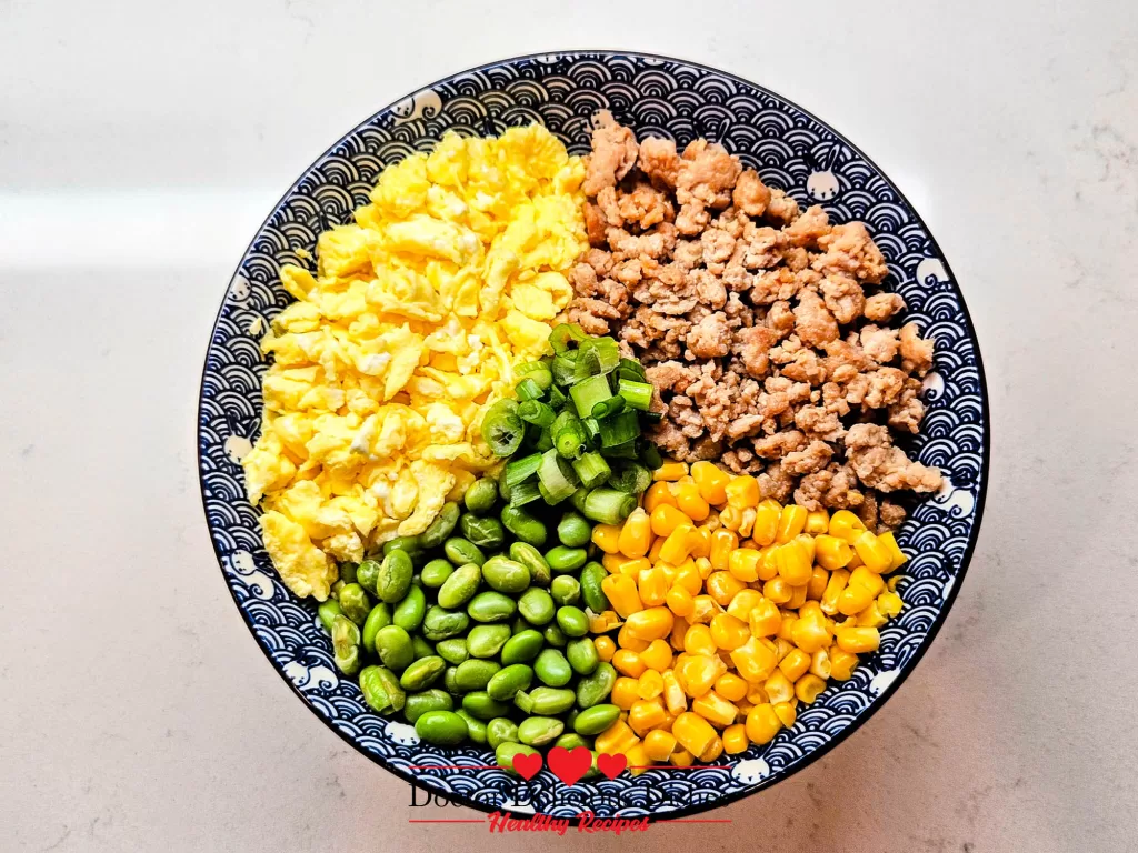 Overhead view of Chicken Soboro Don arranged with ground chicken, fluffy eggs, corn, edamame, and green onions in a blue Japanese bowl - Doctor Delicious Dishes.