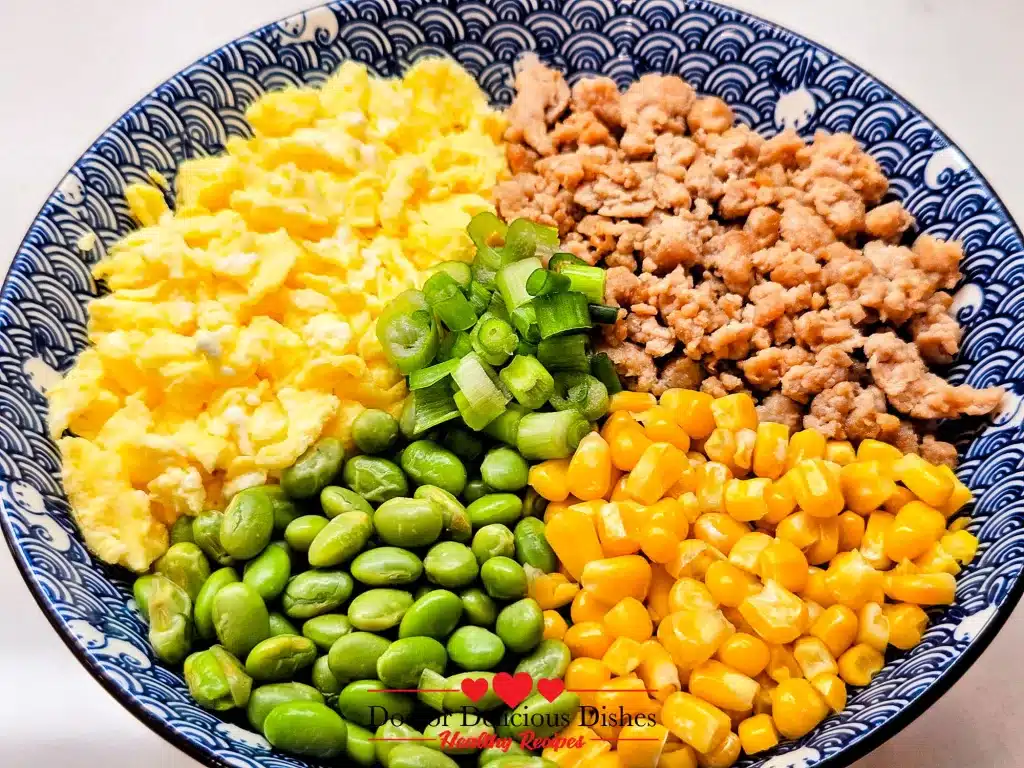Close-up of Chicken Soboro Don showing colorful layers of chicken, eggs, corn, edamame, and green onions in a patterned blue bowl. Doctor Delicious Dishes.