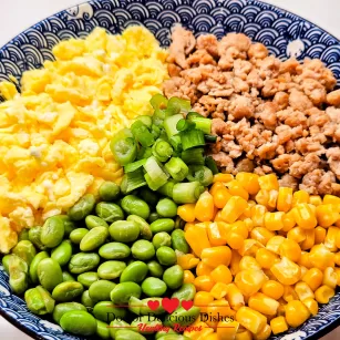 Close-up of Chicken Soboro Don showing colorful layers of chicken, eggs, corn, edamame, and green onions in a patterned blue bowl. Doctor Delicious Dishes.