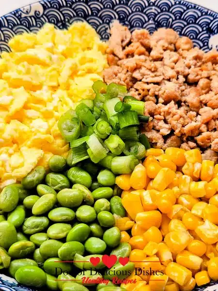Close-up of Chicken Soboro Don showing colorful layers of chicken, eggs, corn, edamame, and green onions in a patterned blue bowl. Doctor Delicious Dishes.