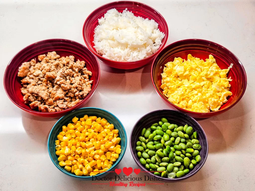 Cooked ingredients for Chicken Soboro Don displayed in colorful bowls with ground chicken, scrambled eggs, rice, corn, and edamame arranged for assembly. Doctor Delicious Dishes.