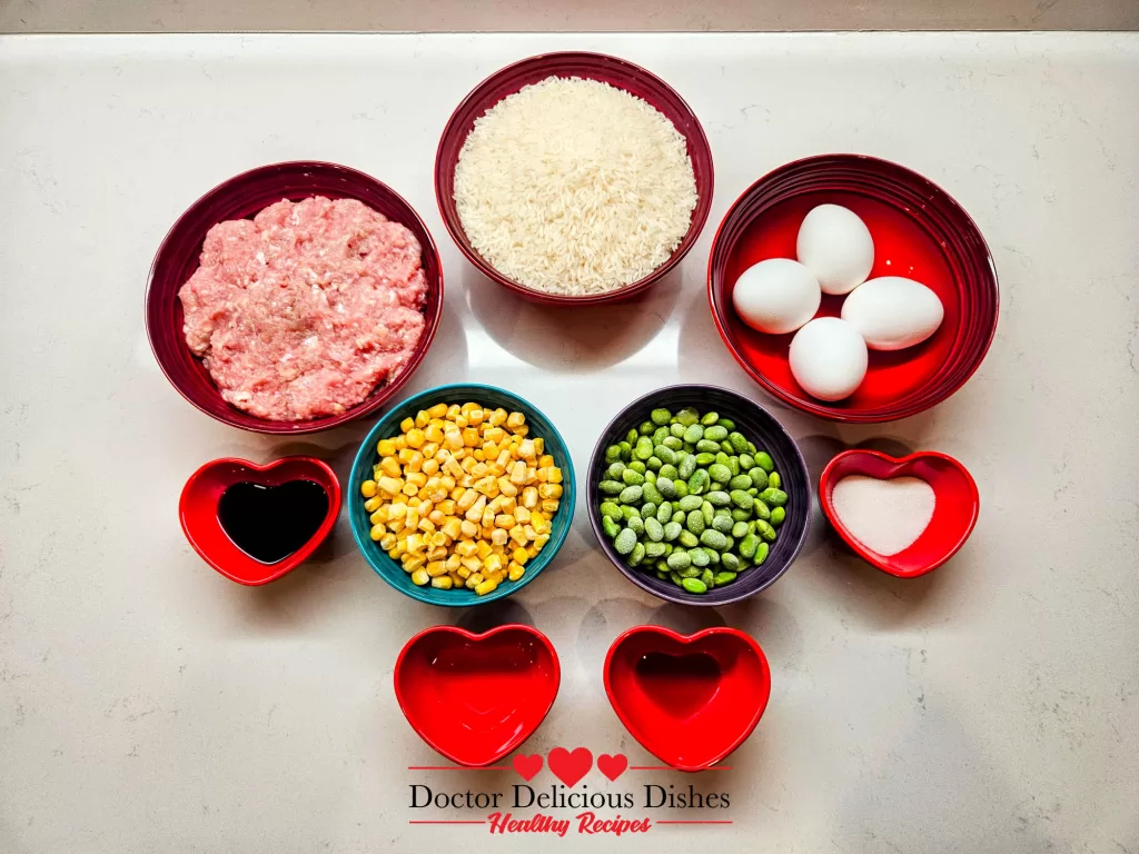 Chicken Soboro Don ingredients arranged in colorful bowls with ground chicken, eggs, rice, edamame, corn, and seasonings on a white counter by Doctor Delicious Dishes.
