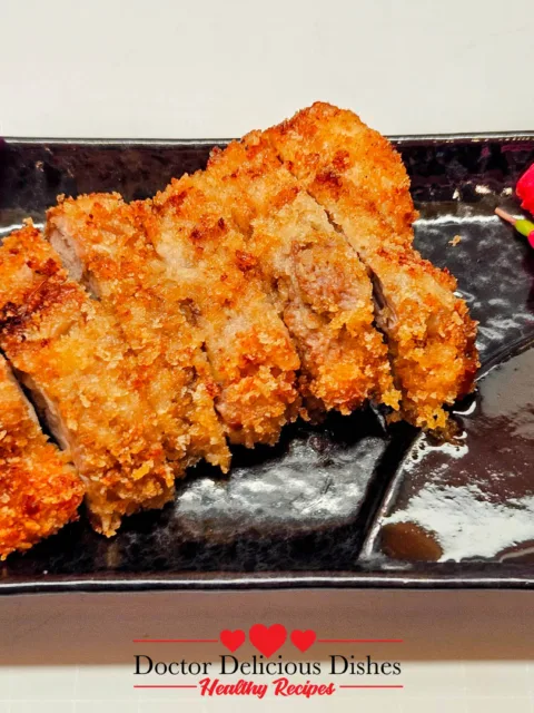 Air fryer chicken katsu recipe sliced and arranged on a black plate with bright edible flowers and dipping sauce.